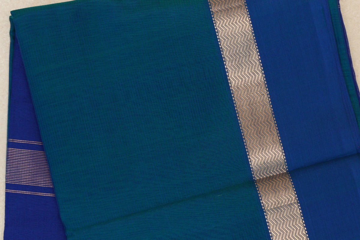 Folded Peacock Blue Maheshwari silk cotton saree showing a subtly checked body, gold-toned zigzag border and deep blue striped fabric peeking out.