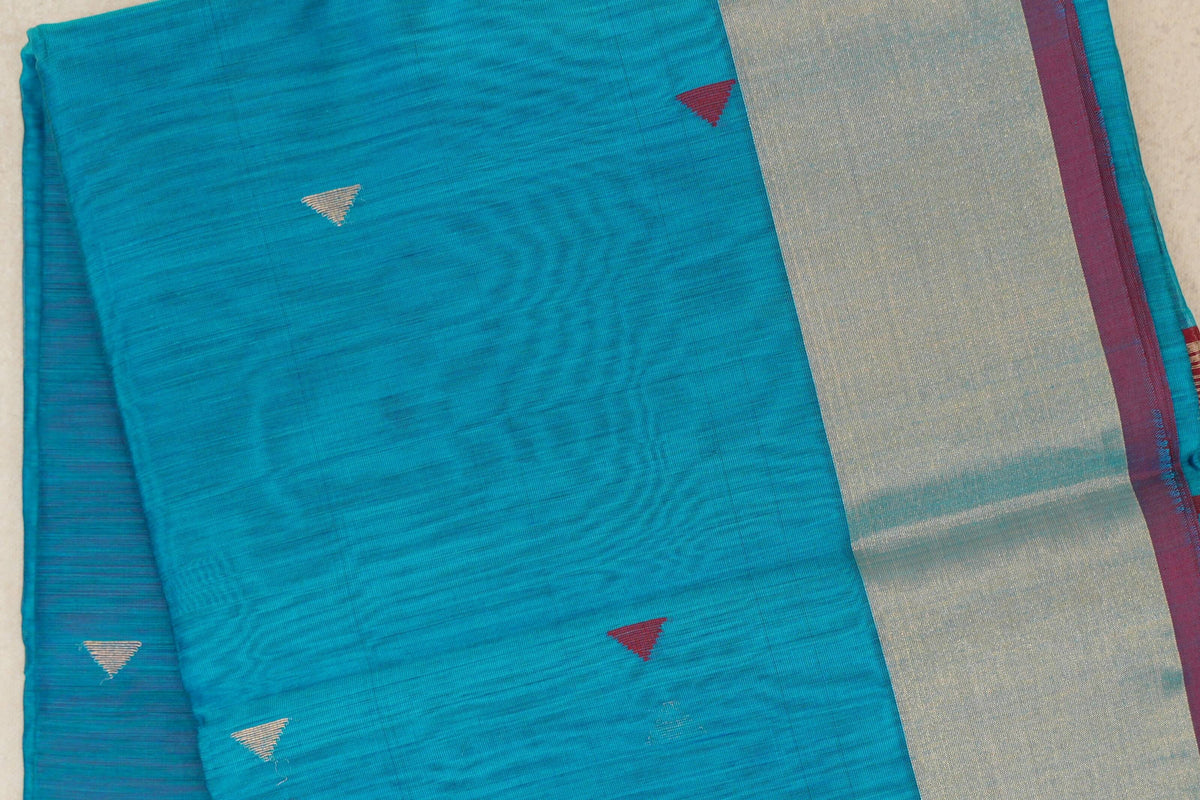 Folded bright turquoise blue silk cotton saree with small red and silver triangular motifs, a broad silver-toned border and deep red edge.