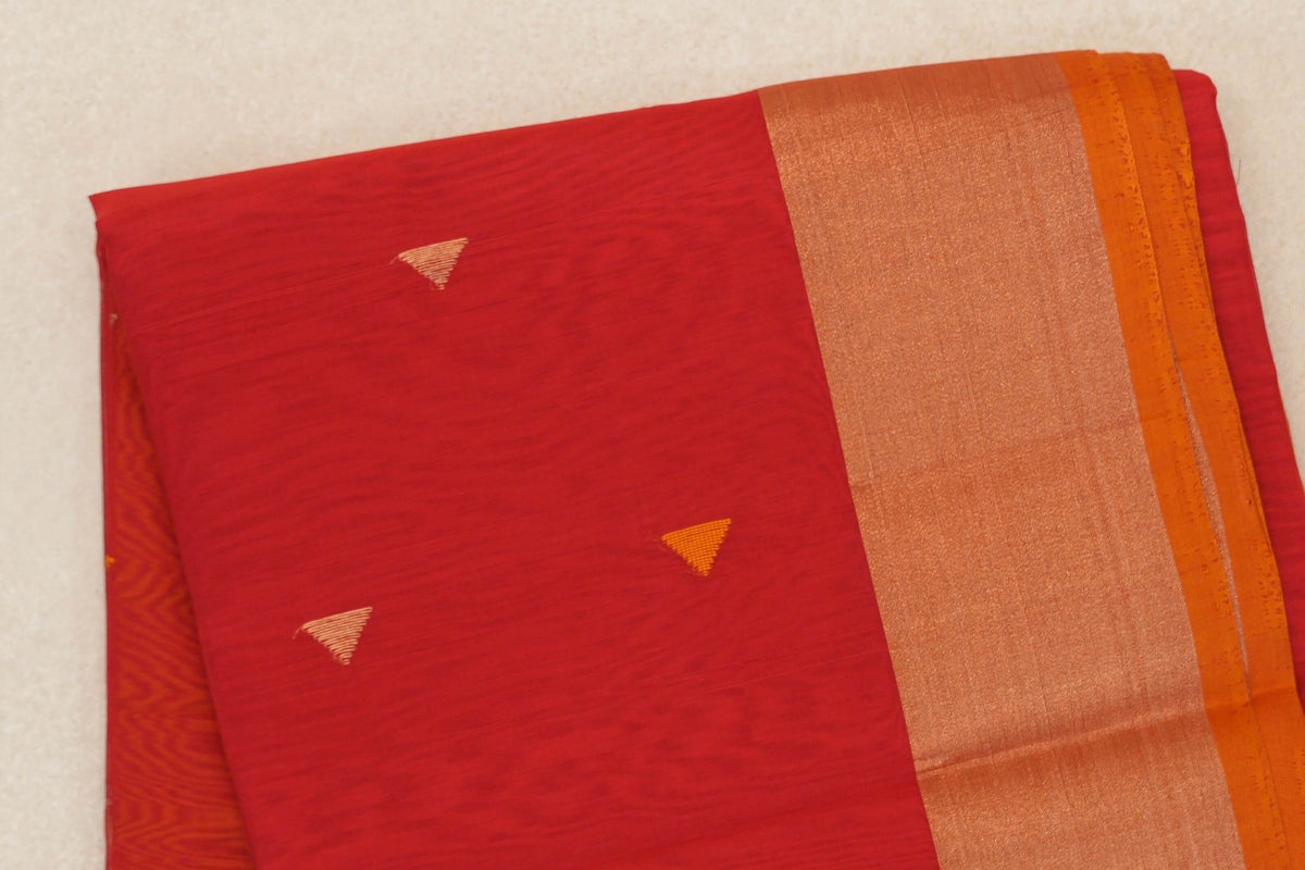 Folded tomato red Maheshwari silk cotton saree with small triangular accents, a broad shimmering border and deep orange selvedge.