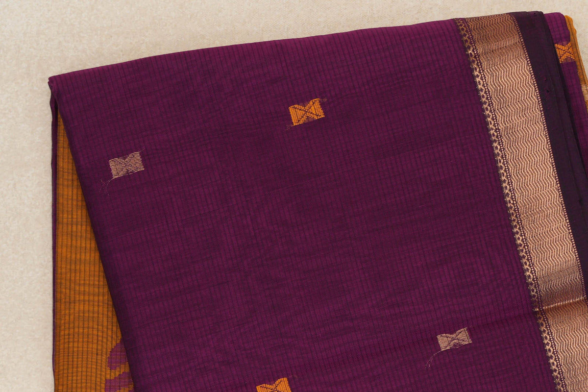 Folded deep magenta silk cotton saree with fine stripes, scattered gold and mustard orange motifs, a patterned gold border and orange fabric peeking out.