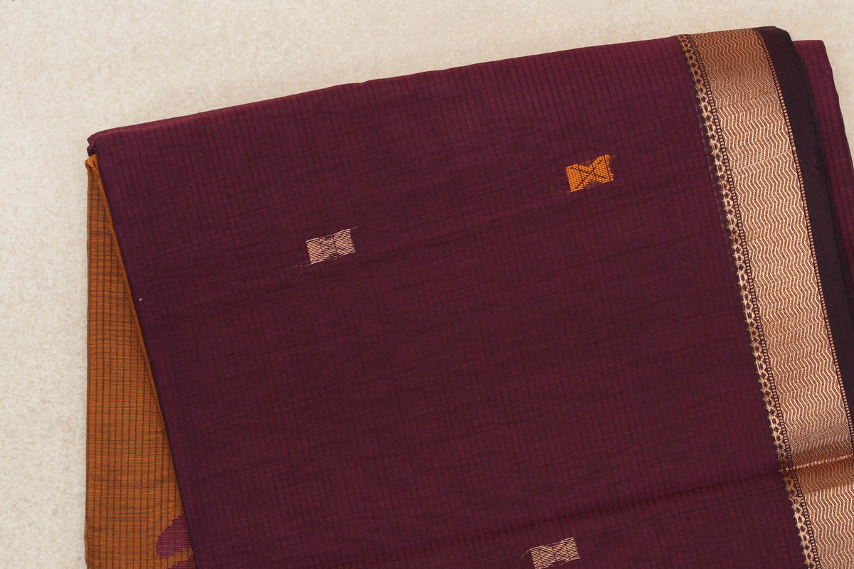 Folded deep wine Maheshwari silk cotton saree with small gold and orange motifs, a gold zigzag border, and deep mustard orange fabric peeking out.