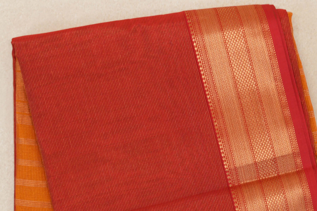 Folded tomato red Maheshwari silk cotton saree with fine stripes and a wide gold-toned geometric border; deep orange fabric peeks out beneath.