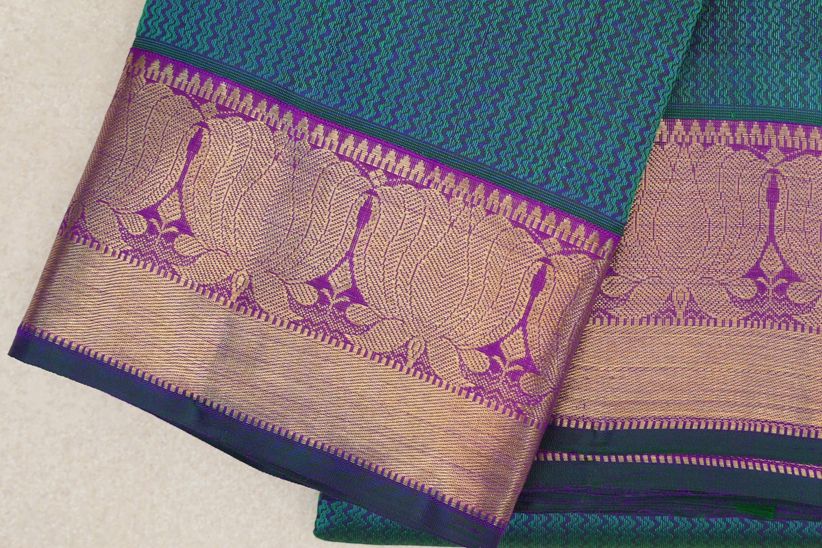 Folded Kanjivaram silk saree showing a patterned peacock green body and broad purple border with gold zari designs and bottle green edging.