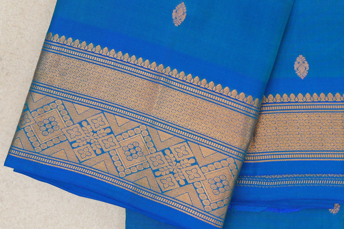 Folded peacock blue lightweight Kanchipuram silk saree with scattered gold motifs and a wide gold zari border with floral and geometric patterns.