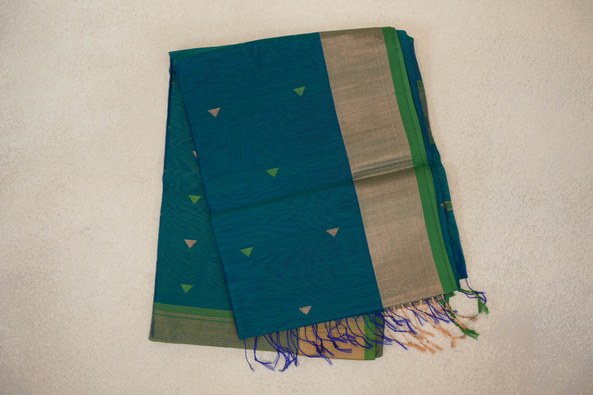 Folded peacock blue Maheshwari silk cotton saree with small triangle motifs, a broad beige border, parrot green edge and colorful tassels.