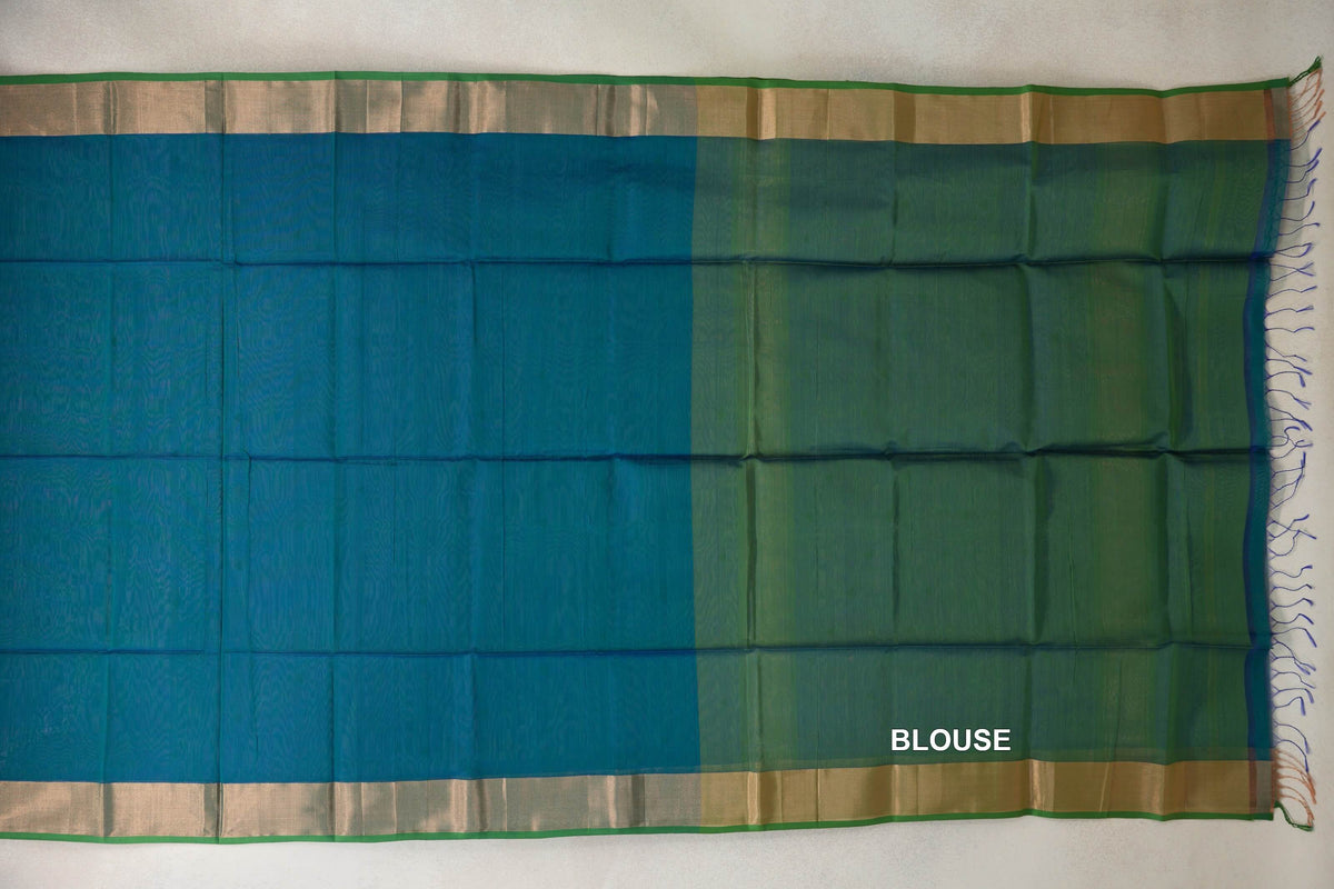 Maheshwari silk cotton saree laid flat, with a peacock blue body, broad shimmering borders edged in green, and a parrot green blouse section at right.