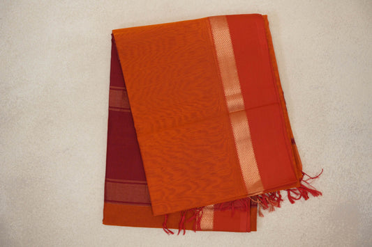 Folded reddish orange Maheshwari silk-cotton saree with an orangish red border, a gold-toned zigzag band, striped fabric and red tassels.