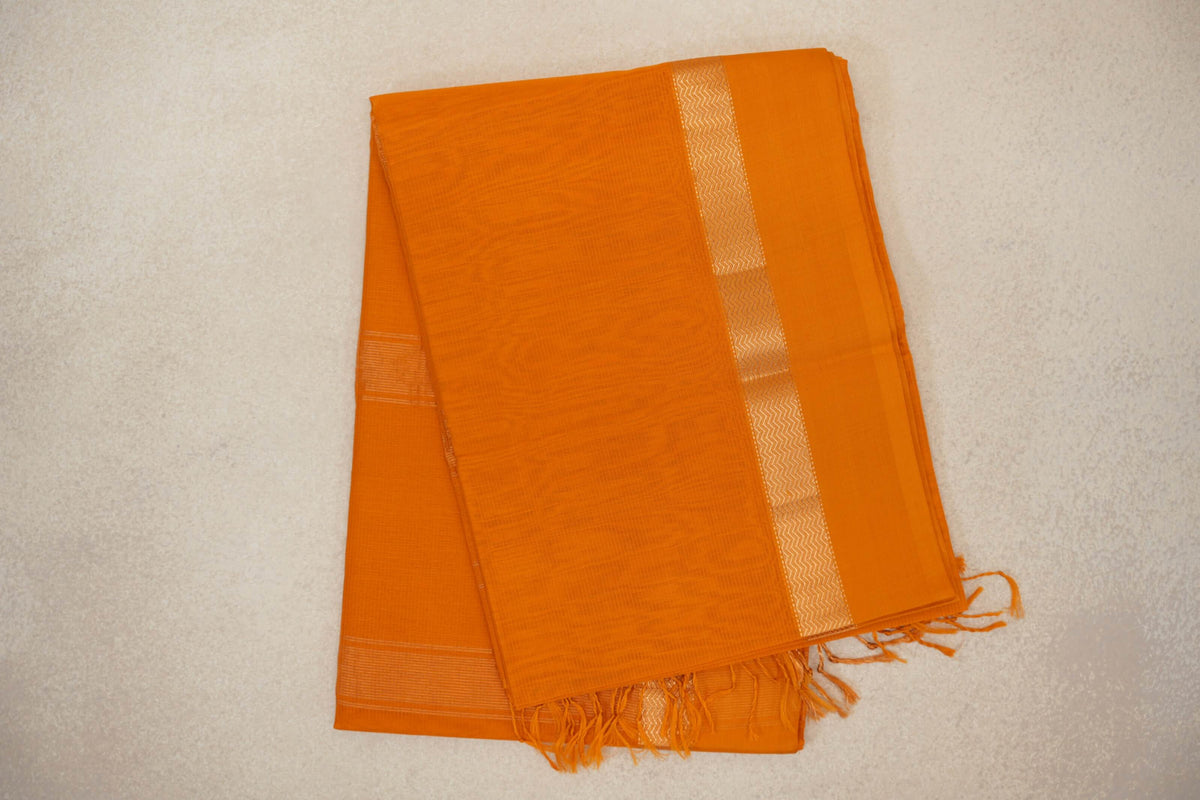 Folded mango yellow Maheshwari silk-cotton saree with a subtle checked weave, a pale zigzag border stripe and tassels at one end.