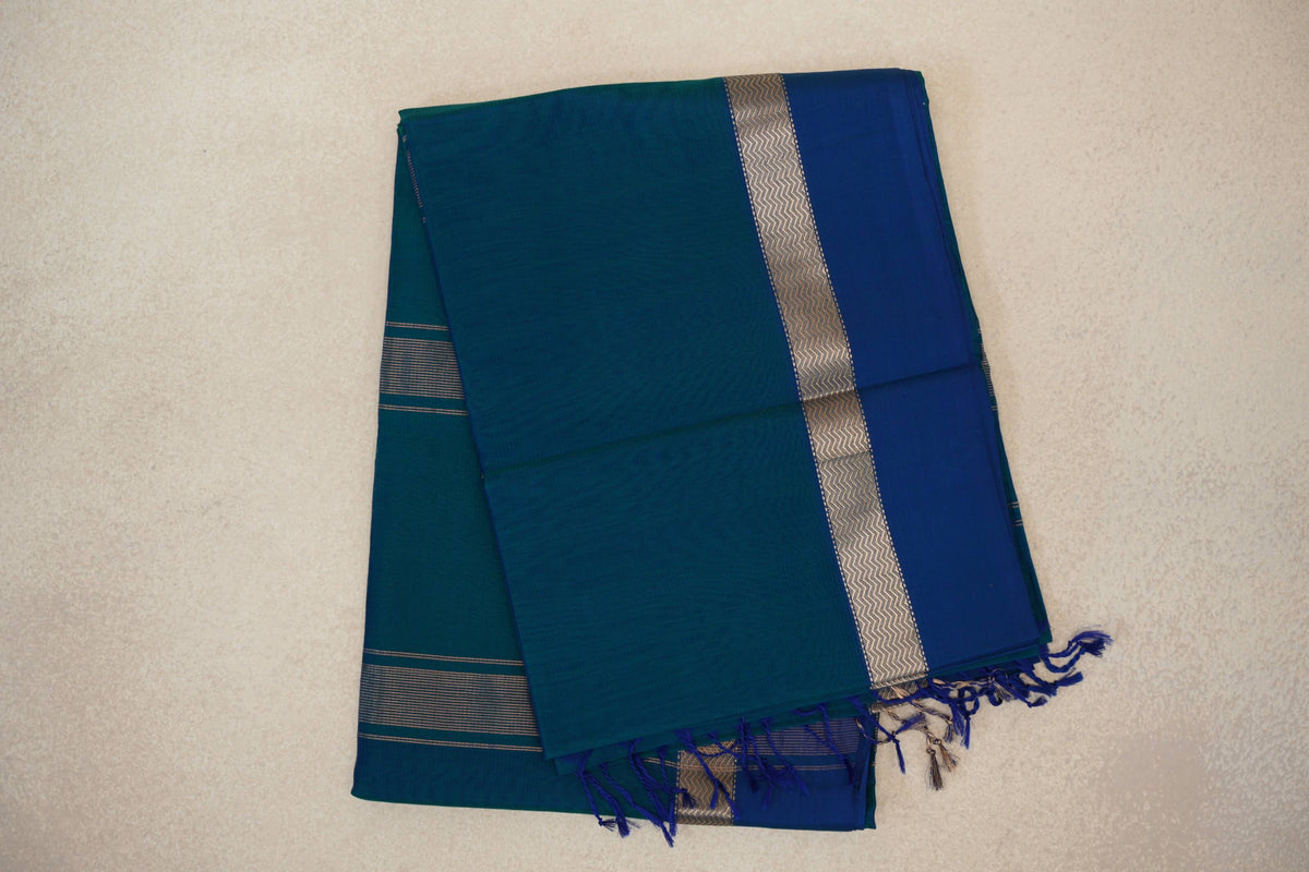 Folded Peacock Blue Maheshwari silk cotton saree showing a plain body, deep Peacock Blue border, metallic zigzag band and tassels.