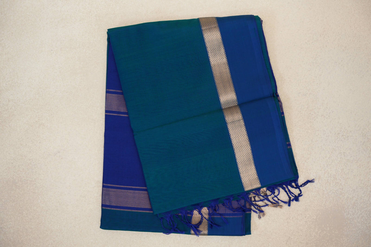 Folded Peacock Blue Maheshwari silk cotton saree with a deep blue border, metallic zigzag band, and a striped pallu with tassels.