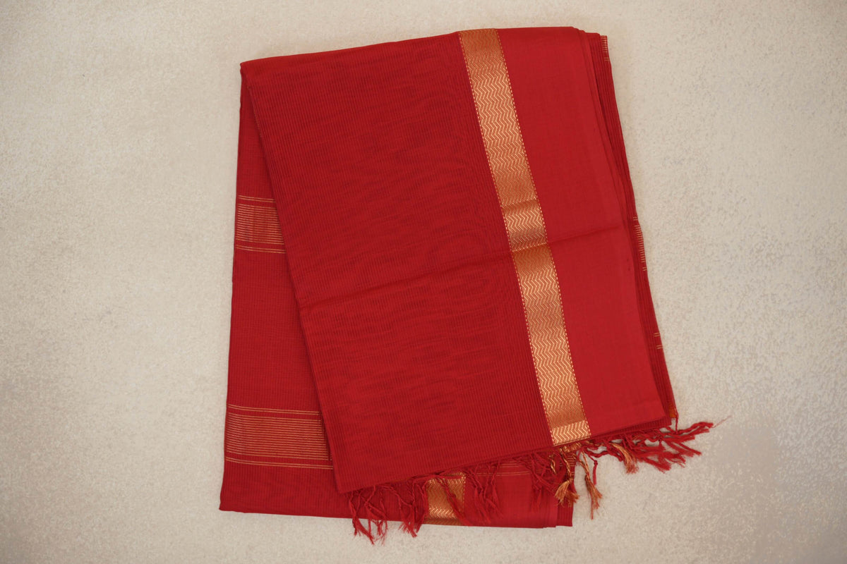 Folded vermilion red Maheshwari silk cotton saree with a fine grid pattern, gold zigzag border, striped detail and tassels.
