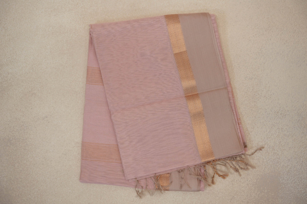 Folded blush pink Maheshwari silk cotton saree with a light sandal beige border, gold-toned zigzag detailing and tassels at one end.