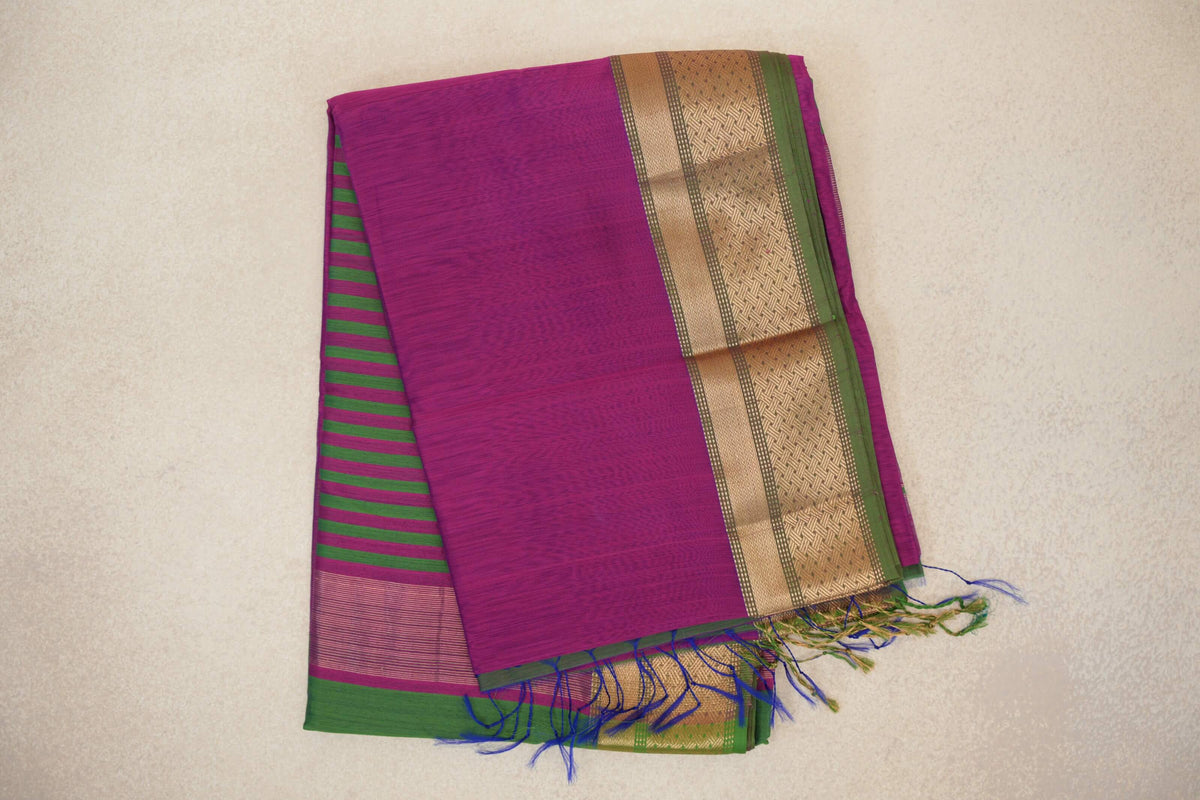 Folded dark magenta silk cotton saree with a gold-patterned border edged in leaf green, green-striped pallu peeking out and loose tassels.