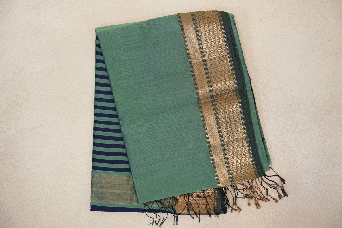 Folded sea green silk cotton saree with a dark sea green and gold zari border, navy-striped pallu peeking out beneath and tassels at the edge.
