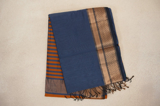 Folded dark grey blue Maheshwari silk cotton saree with a deep orange striped pallu peeking out, a patterned tan border and dark tassels.