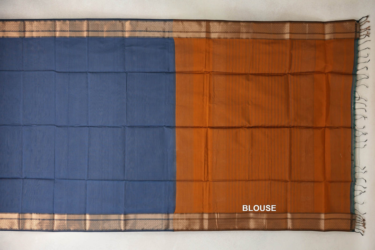 Saree laid flat with a dark grey blue silk cotton body, a deep orange blouse piece, patterned borders and tassels along the right edge.