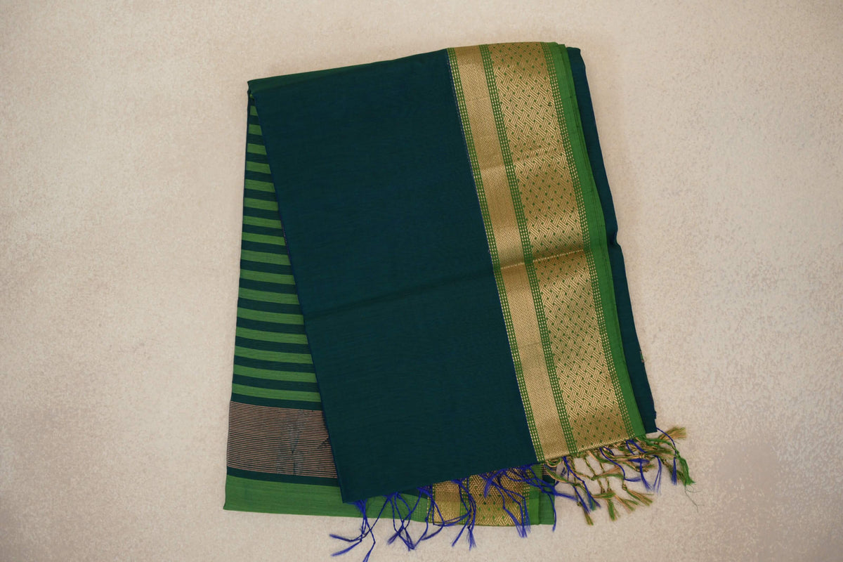 Folded peacock blue Maheshwari silk cotton saree with a leaf green, gold-patterned border, striped pallu peeking out and blue-green tassels.
