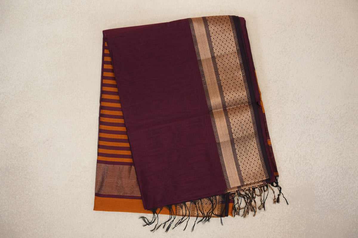 Folded deep plum Maheshwari silk cotton saree with a gold-toned patterned border, deep orange striped pallu peeking out and tassels.