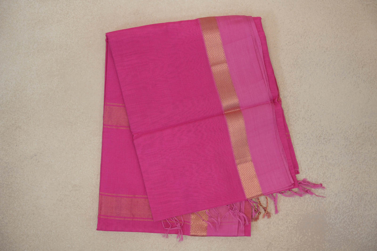 Folded bright Rani Pink Maheshwari silk cotton saree with fine stripes, a pink border, a gold-toned zigzag band and tassels.