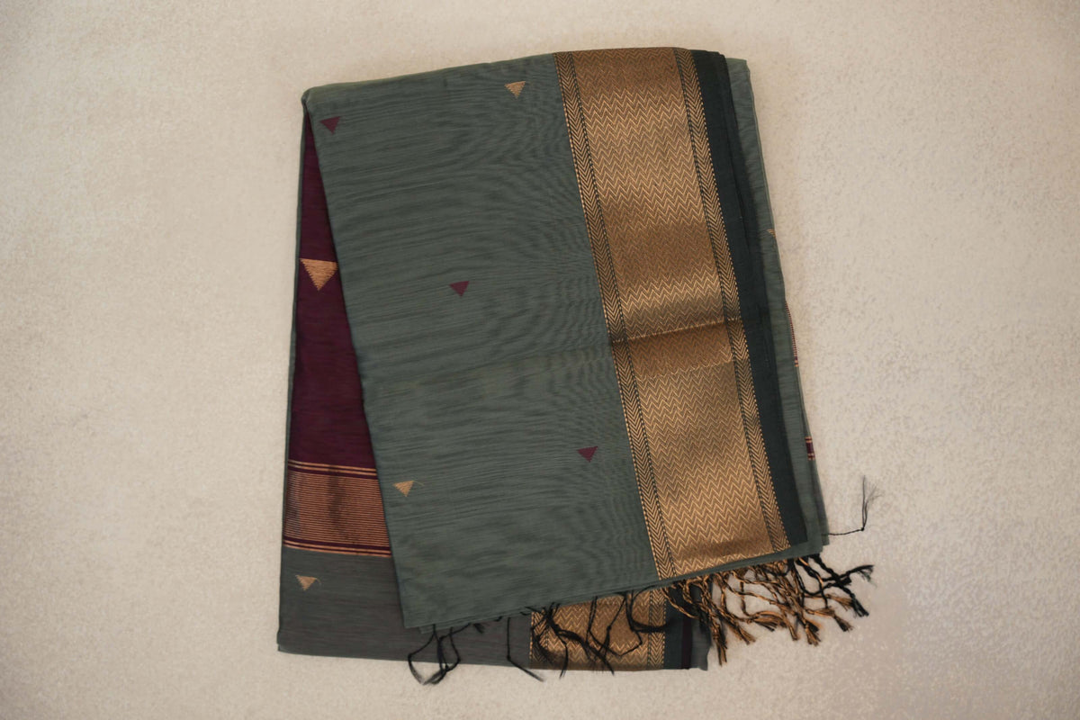 Folded sage green Maheshwari silk cotton saree with small triangle motifs, a dark sage green border with gold chevrons, and a glimpse of the deep wine maroon pallu.