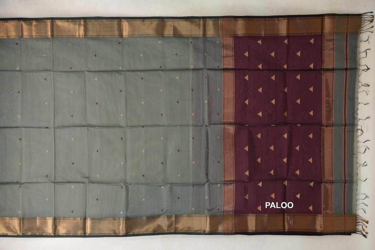 Sage green silk cotton saree laid flat, with small triangular motifs, dark sage green patterned borders and a deep wine maroon pallu with tassels.
