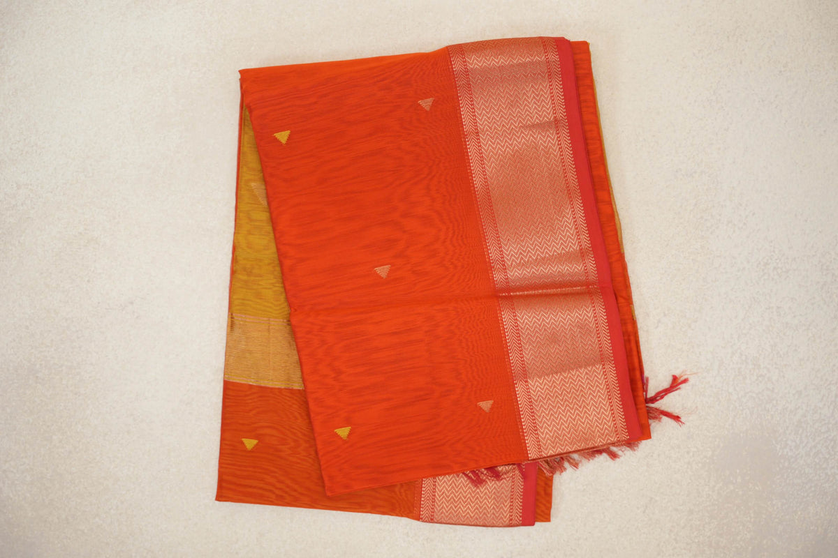 Folded Maheshwari silk cotton saree with a reddish orange body, small triangle motifs, an orangish red chevron border and a mustard yellow section peeking out.
