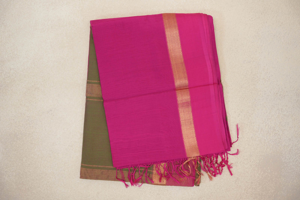 Folded rani pink Maheshwari silk cotton saree with a gold-toned zigzag border and pink tassels; olive green fabric peeks out beneath.