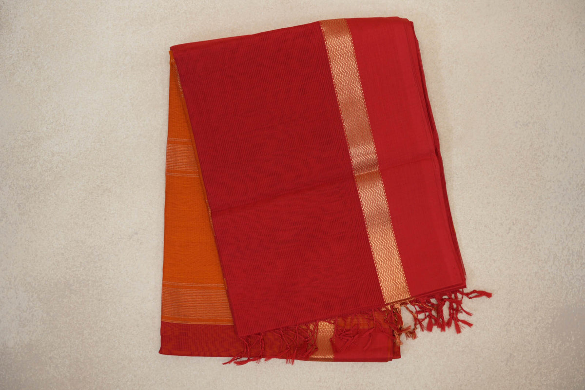 Folded vermilion red Maheshwari silk-cotton saree with a gold-toned zigzag border, deep orange fabric peeking beneath and red tassels.