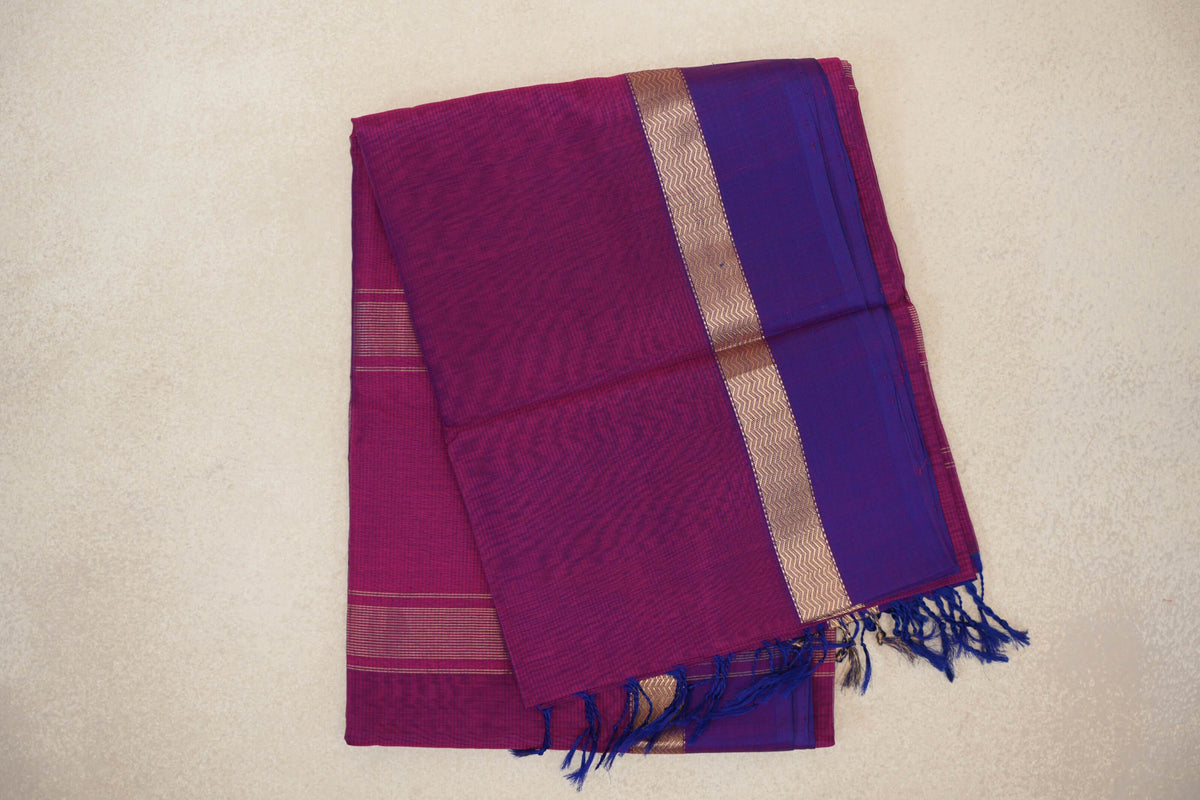 Folded deep magenta Maheshwari silk cotton saree with a violet purple border, gold zigzag band and blue tassels.