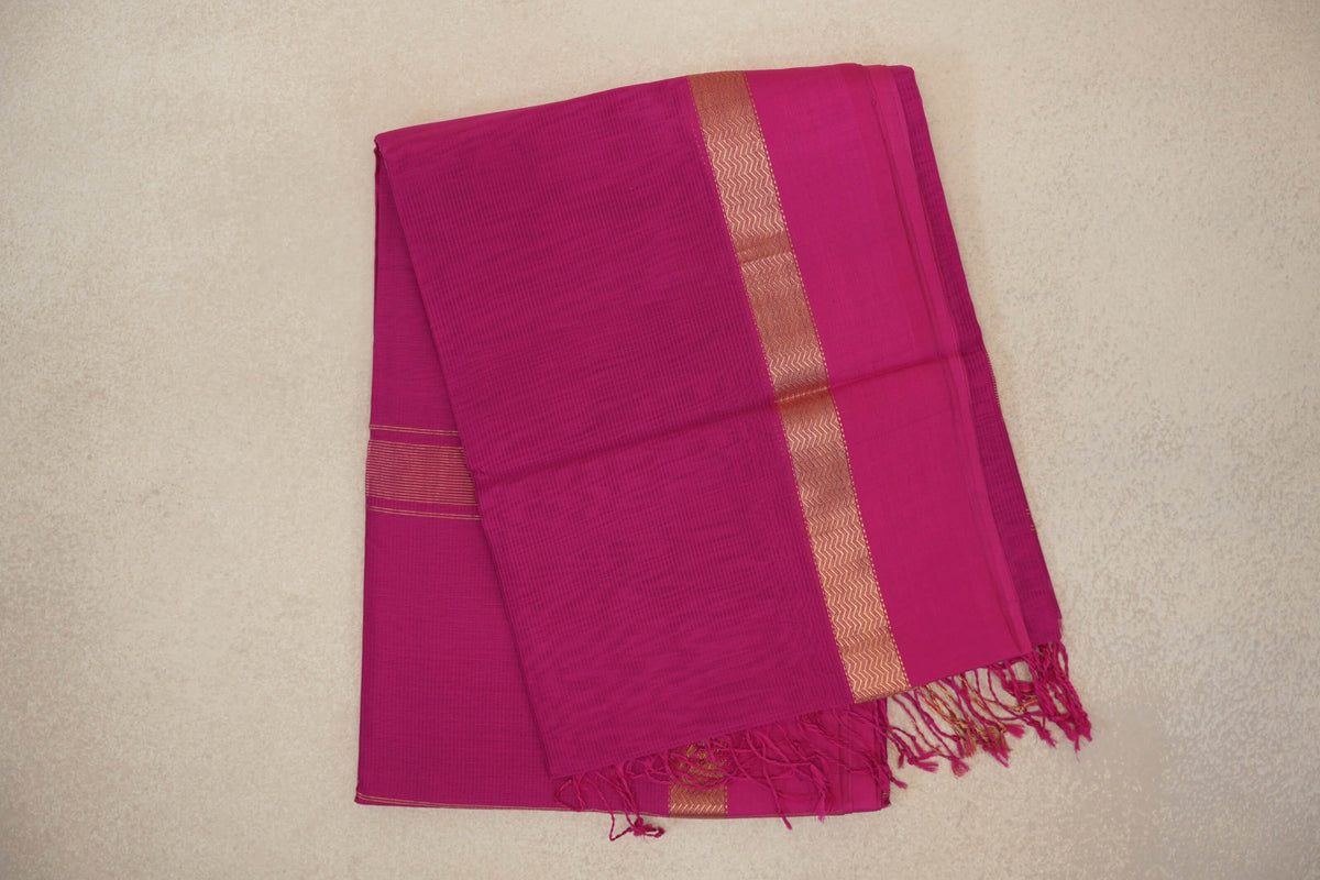 Folded bright rani pink Maheshwari silk cotton saree with a fine checked texture, gold-tone zigzag border and pink tassels.