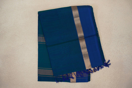 Folded Peacock Blue Maheshwari silk cotton saree with a Deep Peacock Blue border, gold-toned zigzag trim and blue tassels.
