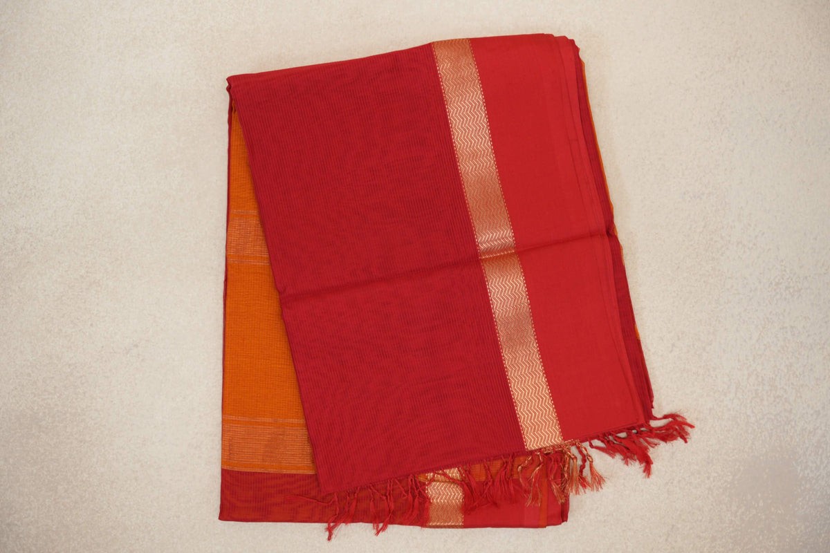 Folded red Maheshwari silk cotton saree with fine stripes, a gold-toned zigzag border, red tassels and orange fabric peeking beneath.