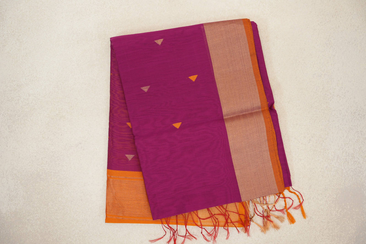 Folded deep magenta Maheshwari silk cotton saree with small triangular motifs, a wide border, orange selvedge and tassels.
