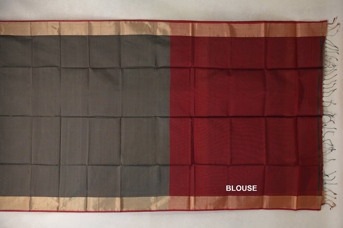 Maheshwari silk-cotton saree laid flat, with a charcoal grey body, deep red blouse section, pale gold-toned borders and tassels at one end.