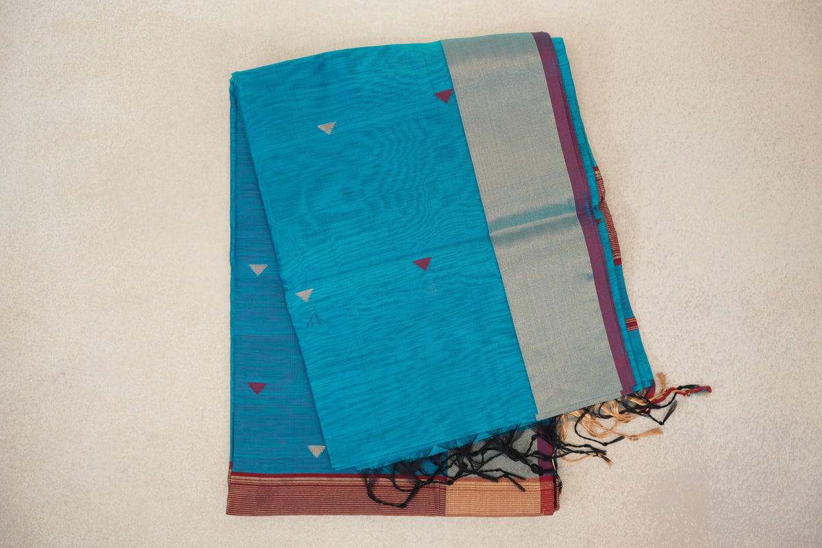 Folded bright turquoise blue silk cotton saree with small triangle motifs, a broad pale metallic border edged in deep red, and tassels.