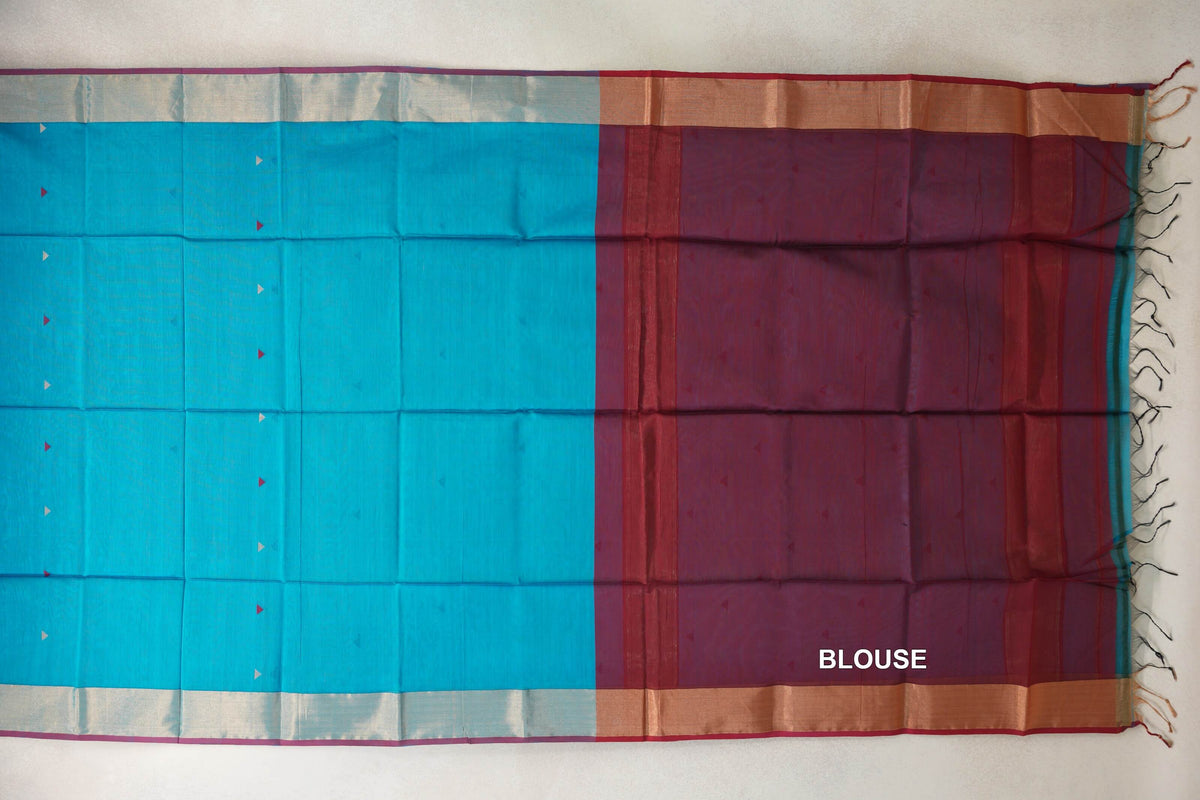 Silk cotton saree laid flat, showing a bright turquoise blue body with tiny triangular motifs, pale borders and a deep red blouse section with tassels.