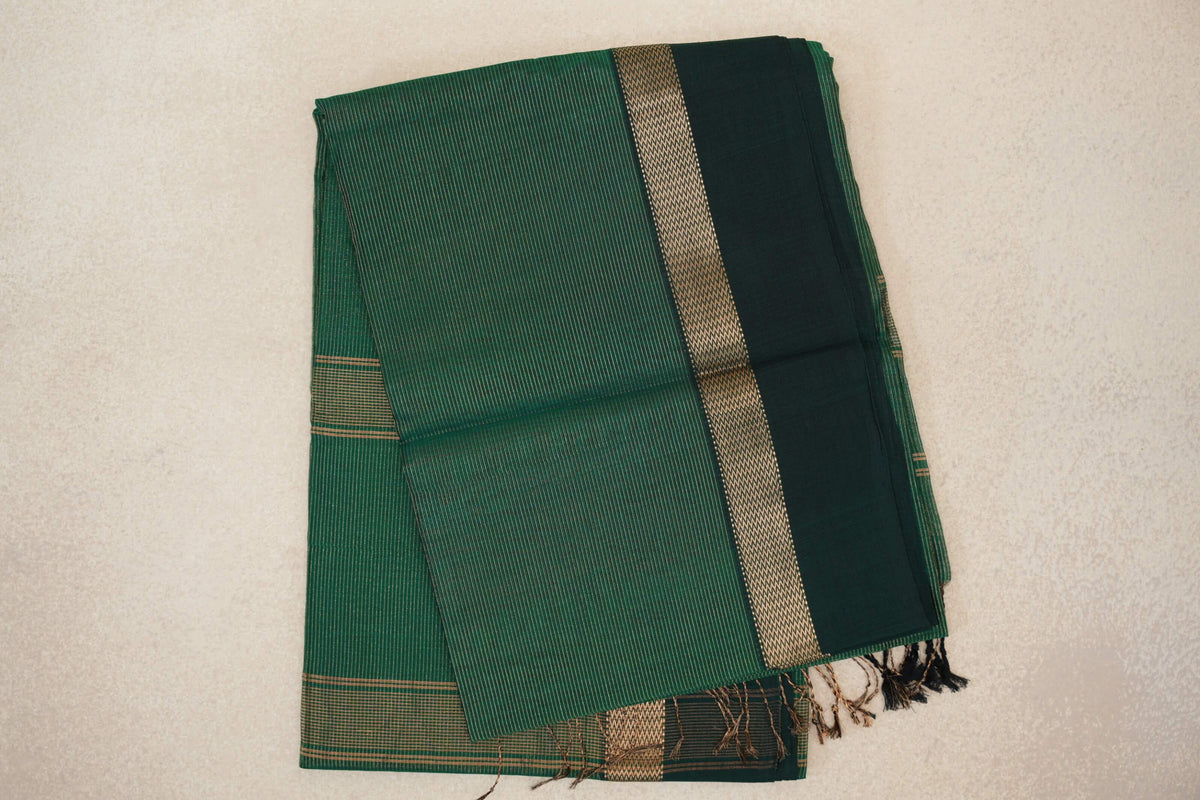 Folded bottle green Maheshwari silk cotton saree with fine stripes, a dark bottle green border, gold zigzag trim and tassels.
