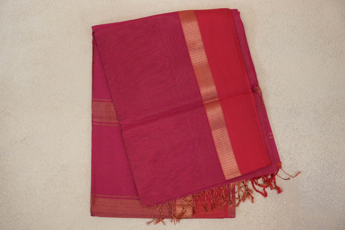 Folded Rani Pink Maheshwari silk cotton saree with fine stripes, a plain panel beside a patterned border, and tassels at the end.