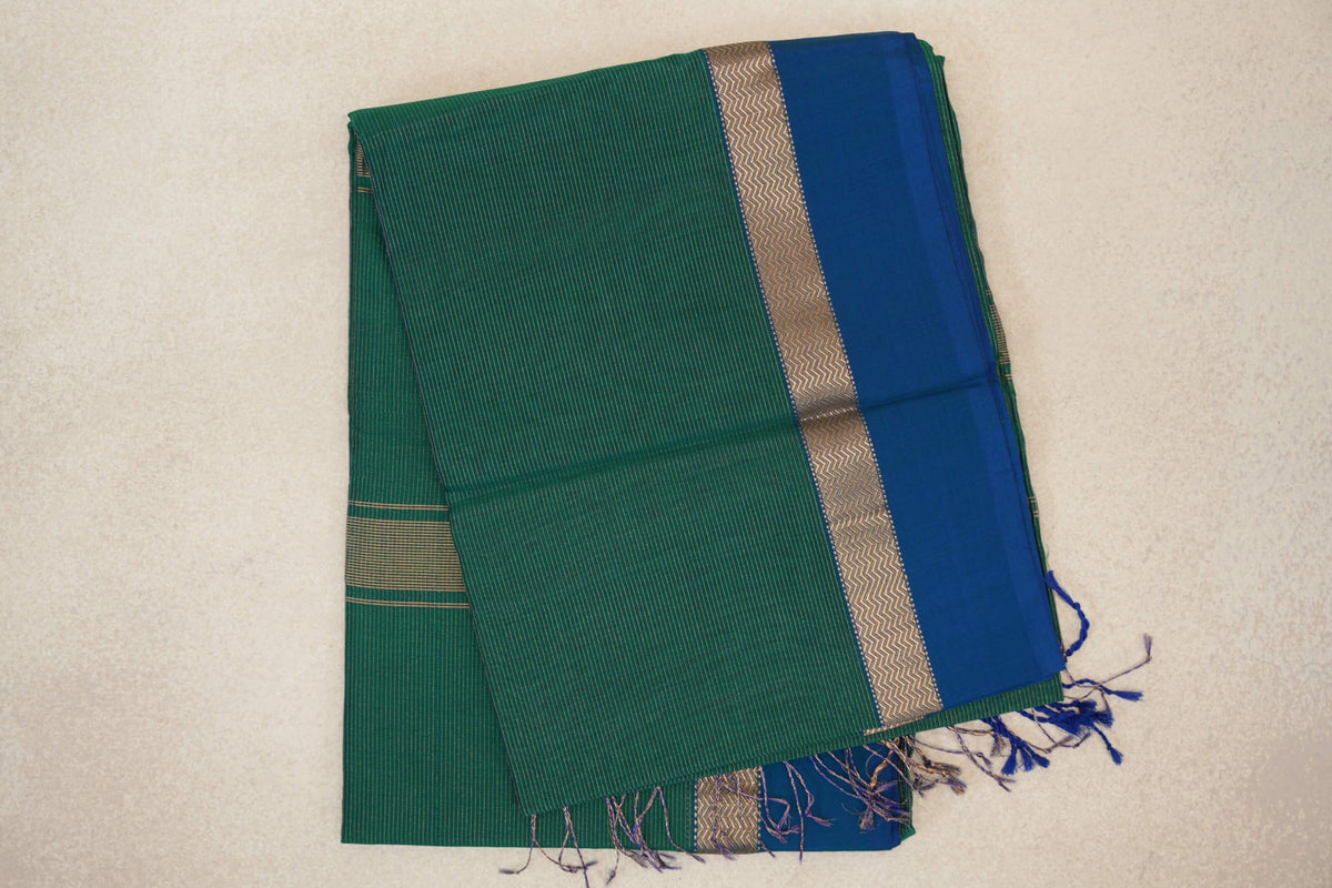 Folded bottle green Maheshwari silk-cotton saree with fine stripes, a peacock blue border, metallic zigzag band and tassels.