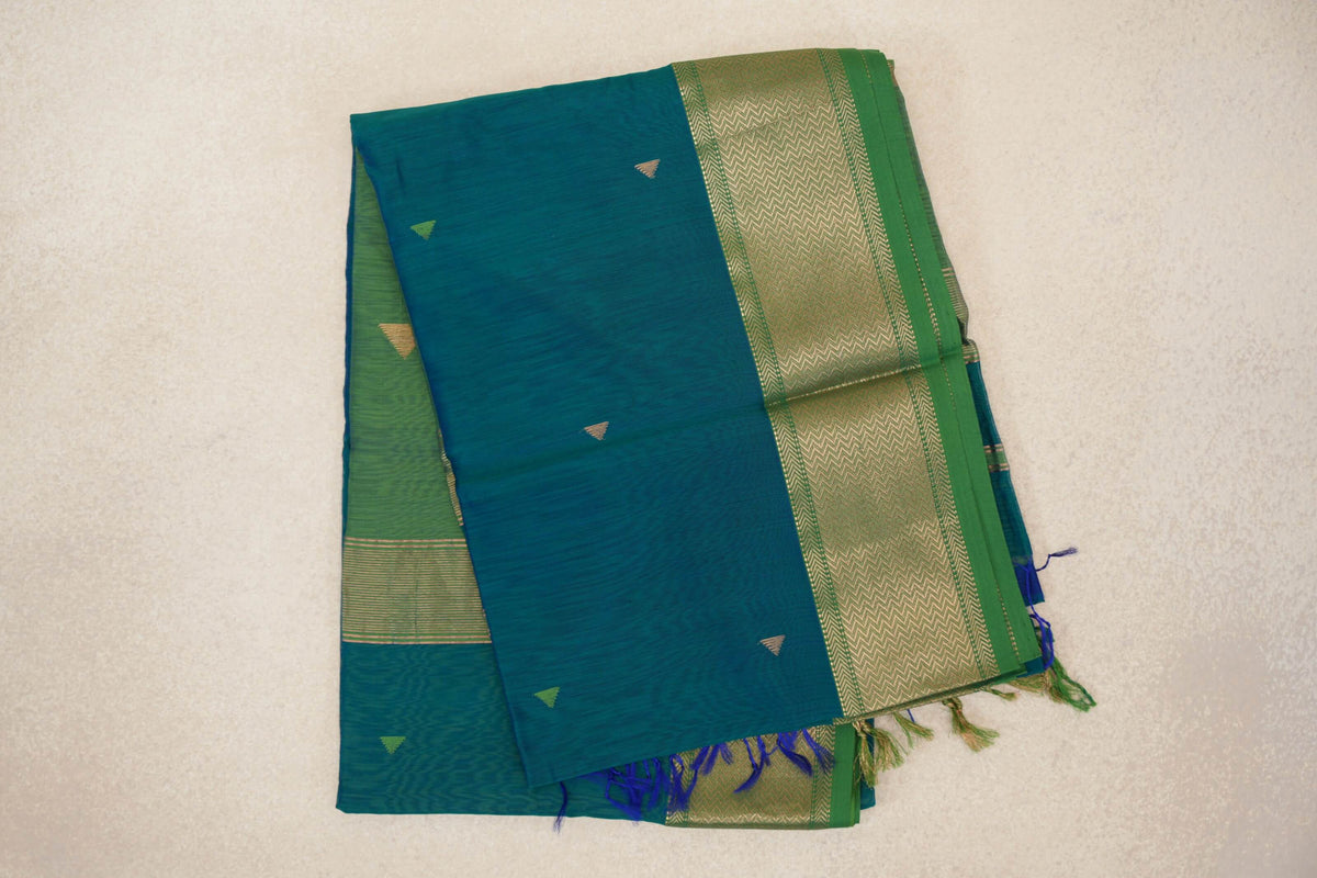 Folded Maheshwari silk-cotton saree with a peacock green body, small triangle motifs, and a leaf green border with gold-toned zigzags and tassels.
