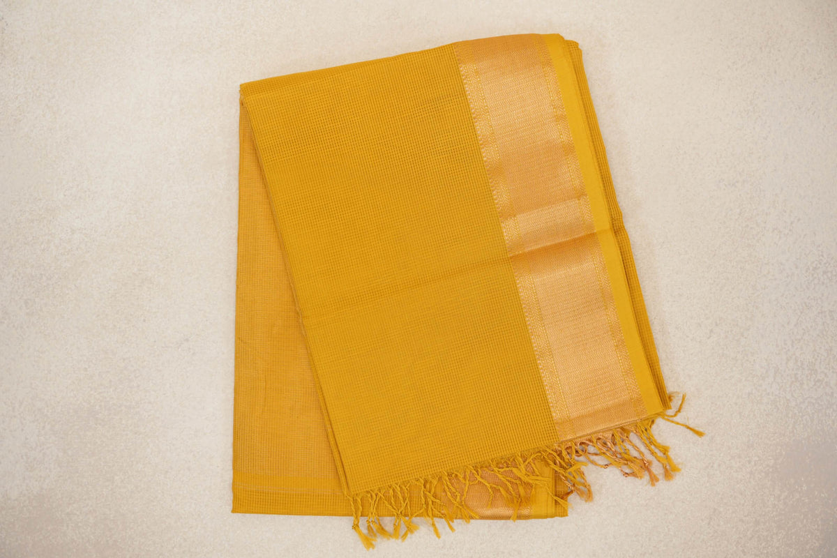Folded mustard yellow Maheshwari silk cotton saree with a fine checked texture, a shimmering woven border and tassels along the edge.
