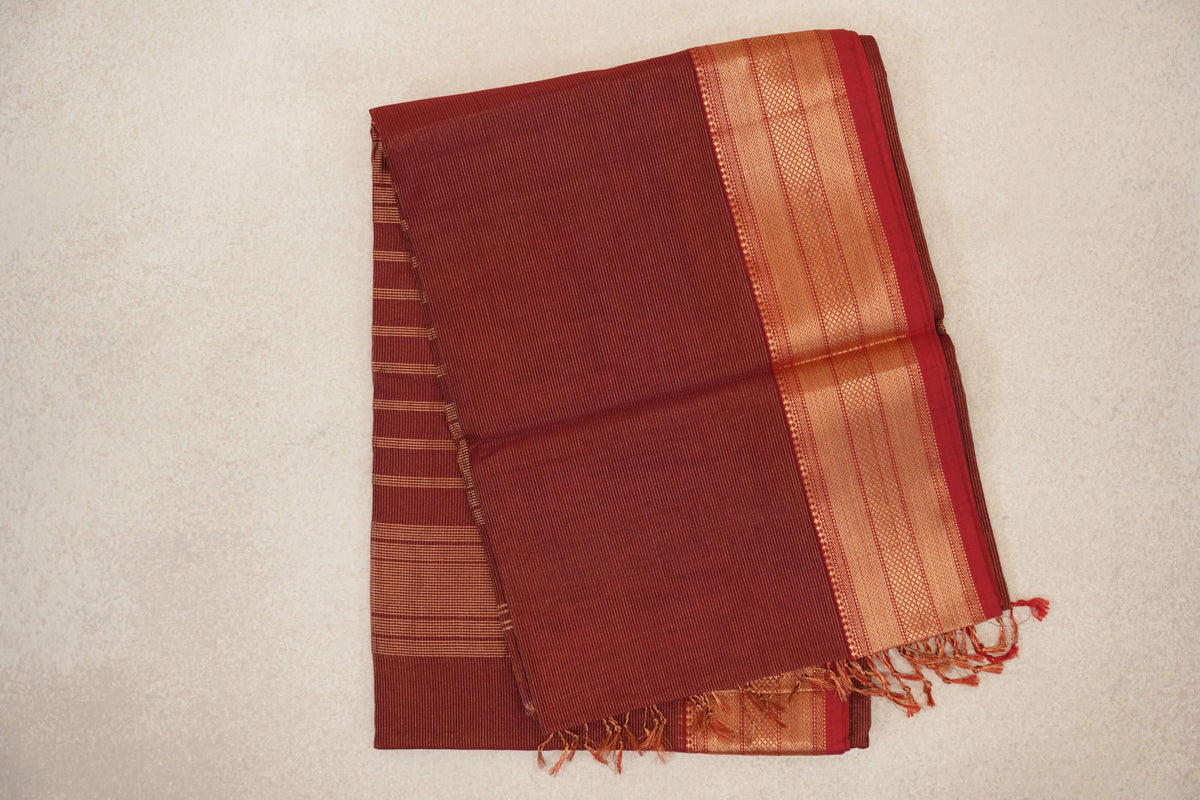 Folded brick red Maheshwari silk cotton saree with fine pinstripes, a patterned gold-toned border and tassels.