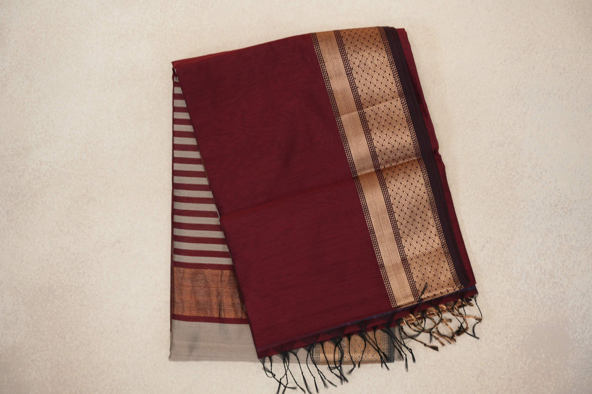 Folded maroon Maheshwari silk cotton saree with a gold-toned patterned border, grey-striped pallu peeking out and dark tassels.