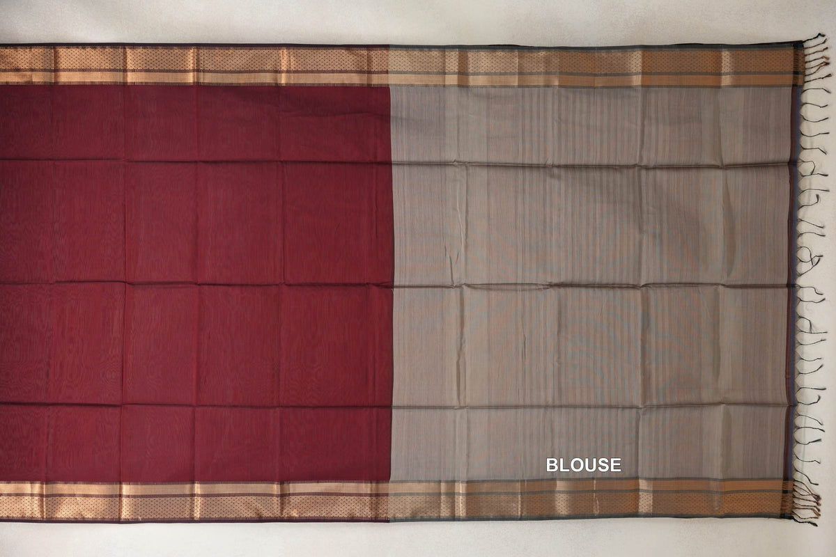Maroon Maheshwari silk cotton saree laid flat beside its grey blouse piece, with patterned copper-toned borders and dark tassels.