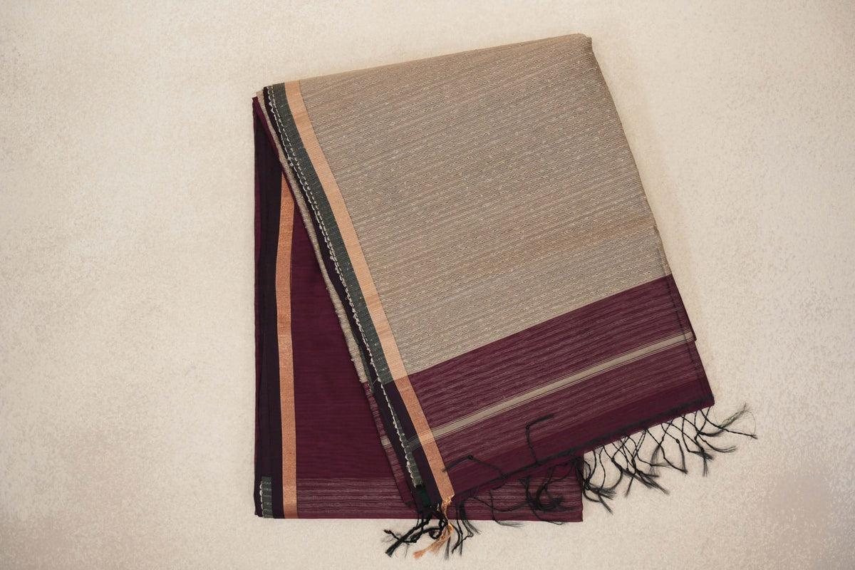 Folded wine maroon Maheshwari silk cotton saree with a sand beige striped pallu, maroon end band and dark tassels.