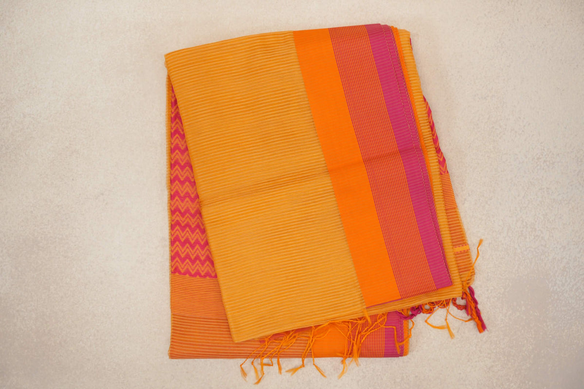 Folded mango yellow Maheshwari silk cotton saree with fine stripes, an orange and rani pink border, a peek of zigzag fabric and tassels.
