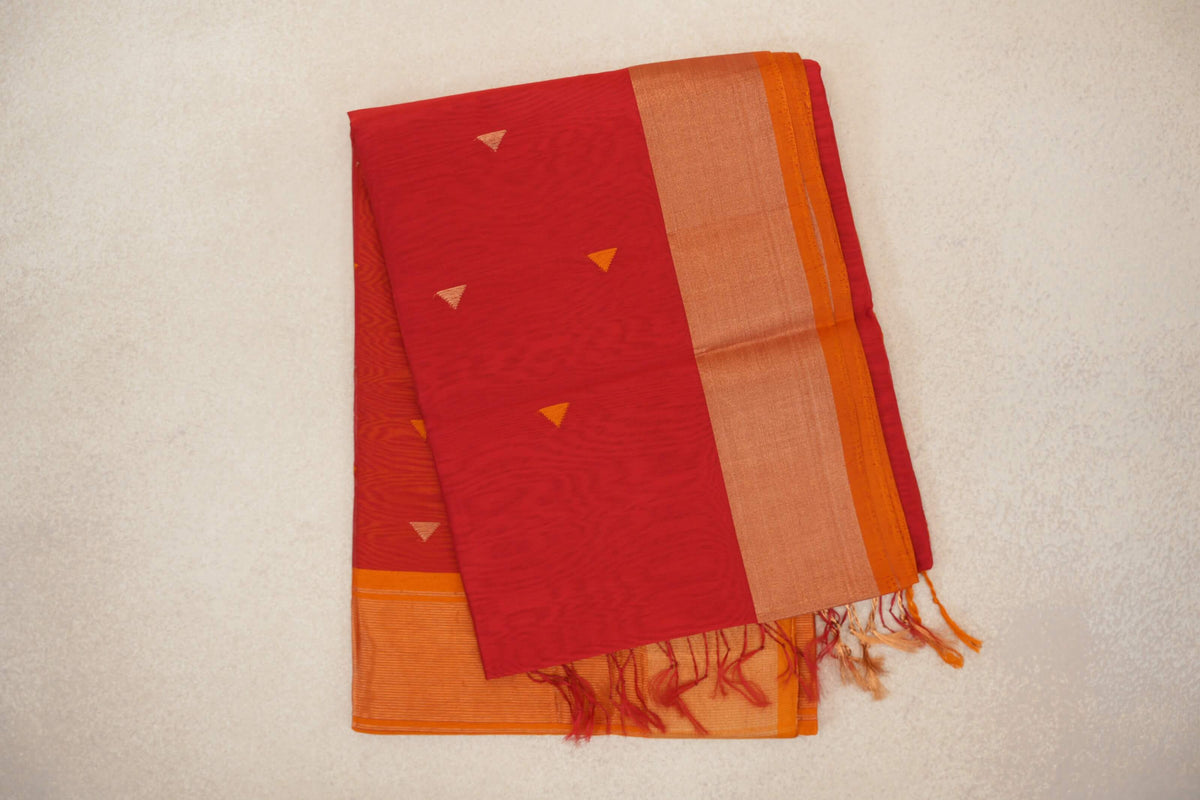 Folded tomato red Maheshwari silk cotton saree with small triangular accents, a broad orange border, deep orange selvedge and tassels.