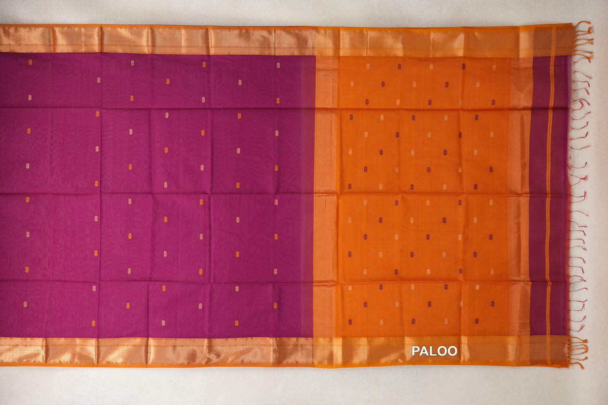 Maheshwari silk cotton saree laid flat, with a dark magenta body, small scattered motifs, deep orange borders and a dotted pallu with tassels.