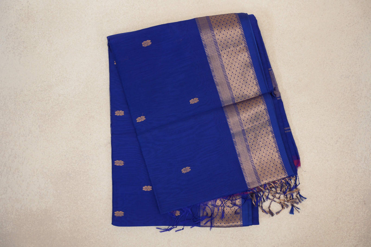 Folded Bright Royal Blue Maheshwari silk cotton saree with small gold-toned motifs, a patterned border and tassels.