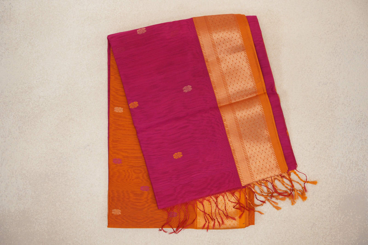 Folded rani pink Maheshwari silk cotton saree with small floral motifs, a deep orange woven border, and an orange pallu with tassels peeking out.